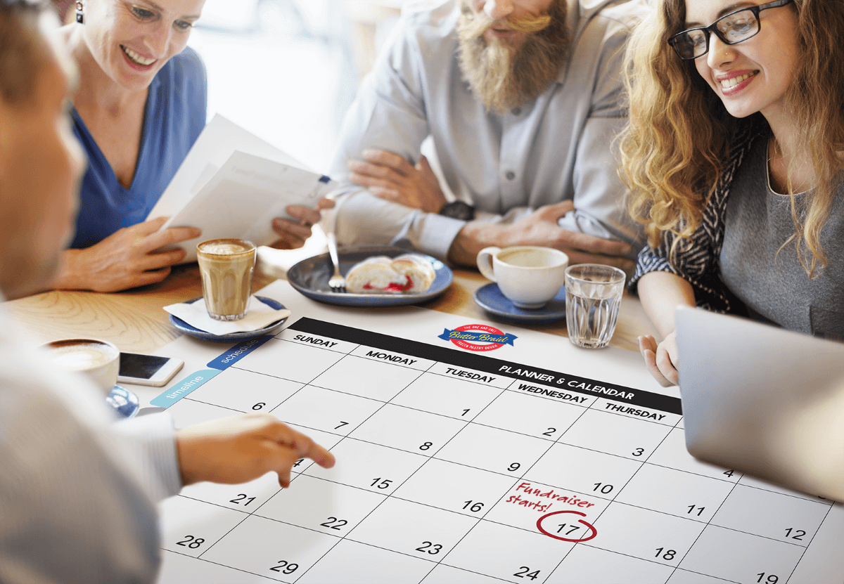 Group of people around a large calendar with dates circled, a plate with a pastry slice, and coffees.